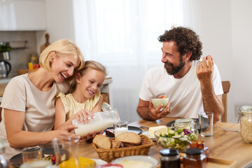 A family shares a joyful breakfast at home, with a mother pouring milk, a daughter smiling, and a...