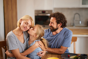 A mother receives a loving kiss from her daughter while the father smiles joyfully. They sit...