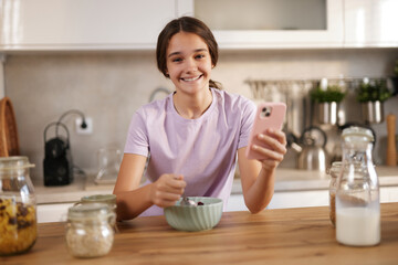 A teenage girl smiles as she sits at a wooden table in a bright kitchen, eating from a bowl while...
