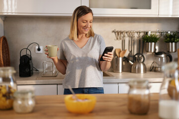 Young woman enjoys her morning routine in a cozy kitchen, sipping coffee while checking her...