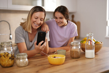 Two smiling women share a moment over breakfast, examining a smartphone in a well-lit kitchen. They...