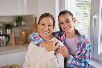 Mother and daughter smile brightly as they enjoy quality time in a well-lit kitchen, sharing a warm...