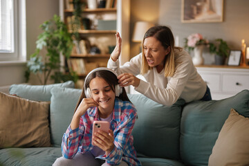 In a comfortable living room, a teenager enjoys music through headphones while her parent shows...