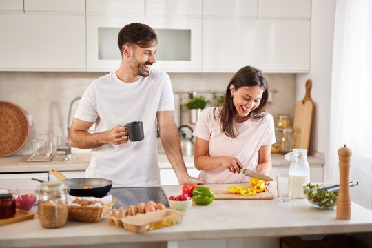 In a modern kitchen, a couple shares laughter while cooking together. The man holds a coffee mug, and the woman chops vibrant vegetables. Sunlight fills the space, enhancing the warmth.
