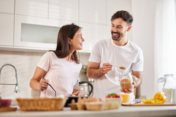 A happy couple is preparing breakfast in a bright kitchen. They are smiling at each other while measuring ingredients, surrounded by fresh fruit and cooking utensils.