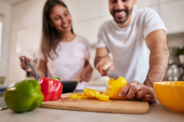 A couple happily prepares a meal in their modern kitchen, chopping vibrant yellow and red peppers, sharing smiles and teamwork while enjoying their time together.