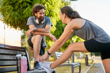 Two individuals are engaged in a fitness activity, with one stretching while sitting on a park...