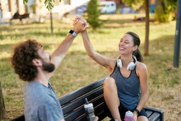 A man and woman high-five each other after a workout session in a park, both smiling while seated...
