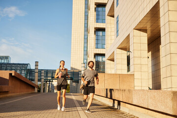 Two individuals run side by side along a spacious pathway surrounded by modern buildings. They are...