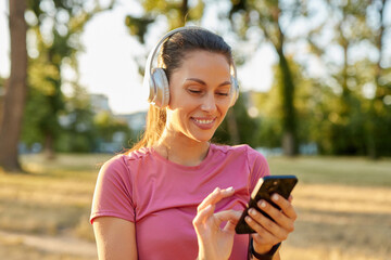 A woman in athletic attire smiles while using a smartphone in a park. She wears headphones and...