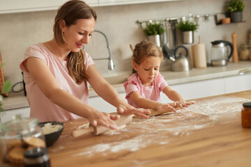 A mother and daughter joyfully roll out dough in a sunny kitchen. Flour dust fills the air as they...