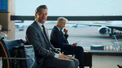 Confident businessman in a suit sits relaxed at the airport terminal, exuding charm and success...