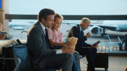 Business dad spends a heartfelt moment with his daughter at the airport, playing with her teddy...