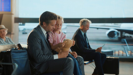 Business dad spends a heartfelt moment with his daughter at the airport, playing with her teddy...