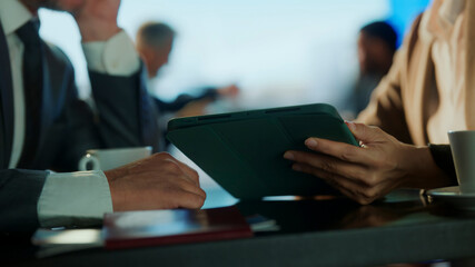 Close-up of hands holding digital tablet during business meeting at airport café, with coffee on the table and blurred professionals in background — symbolizing mobile work, collaboration, and travel  © Videophilia