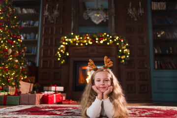 Merry Christmas. Little girl smiling near Christmas tree in classical dark interior. Young happy kid wearing deer horns in living room with fireplace Christmas tree gift boxes. Christmas eve at home