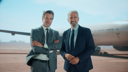 Two confident businessmen in suits stand together in front of a cargo aircraft on the airport...