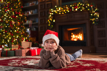 Merry Christmas. Little boy smiling near Christmas tree in classical dark interior. Young happy kid wearing Santa Hat in living room with fireplace Christmas tree gift boxes. Christmas eve at home
