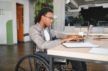 Focused african american employee using laptop in wheelchair at office desk