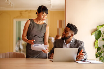 Young businesswoman discussing documents with male professional working over laptop at desk in...