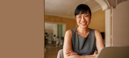 Portrait of female professional with laptop on desk sitting against wall in corporate workplace