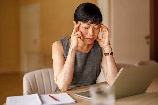 Portrait of stressed businesswoman touching head in pain while working online over laptop at corporate desk