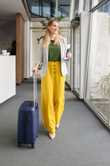 Successful female professional with passport and ticket pulling suitcase while walking in airport lobby