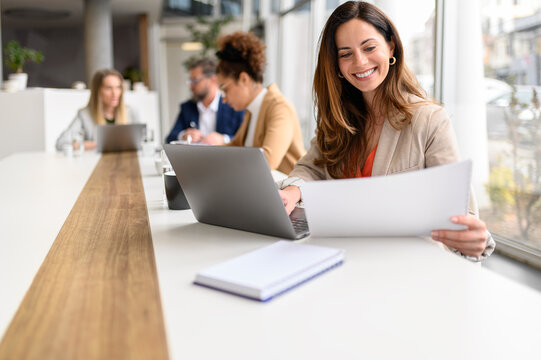 Smiling professional analyzing legal document while working on laptop in modern office with coworkers in the background