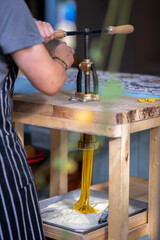 A person operates a manual pasta machine on a wooden table, extruding fresh noodles into a flour-filled tray