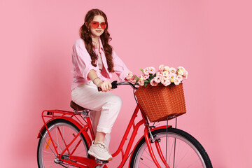 Beautiful teenage girl in sunglasses riding bicycle with basket of flowers on pink background