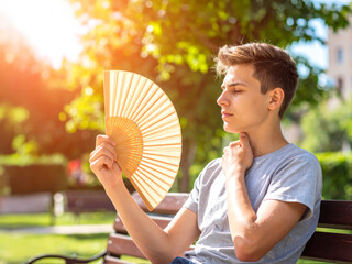 A white male teenager sitting on a park bench during a heatwave, using a handheld fan to cool off