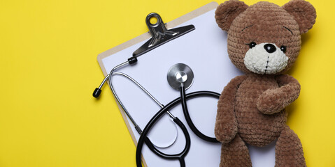 Pediatrics concept. Toy bear with stethoscope and clipboard on yellow background, top view. Space for text