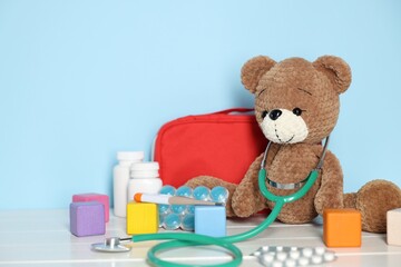 Pediatrician concept. Toy bear, cubes, first aid kit, pills and stethoscope on white wooden table against light blue background © New Africa