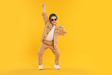 Happy little boy in sunglasses and baseball hat dancing on orange background