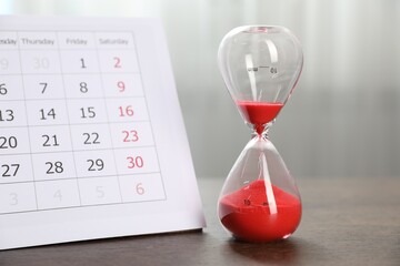 Deadline. Hourglass with flowing sand and calendar on wooden table indoors, closeup