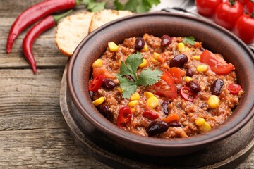 Tasty chili con carne in bowl, ingredients and bread on wooden table, closeup