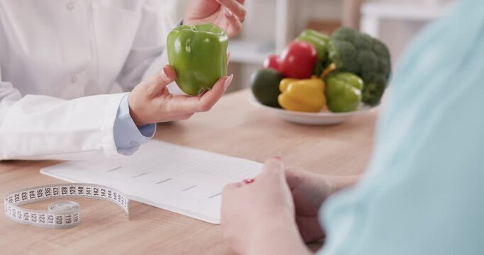 Nutritionist holds green pepper and gives recommendations to patient about healthy diet. On table there are vegetables, symbolizing balanced diet, and centimeter tape, symbolizing monitoring