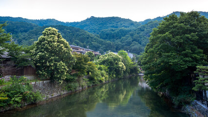 A calm river flows through a quiet district in Kyoto, Japan, bordered by stone walls, blooming trees, and traditional houses. 