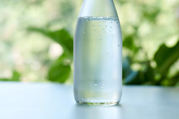 Close-Up of Frosted Glass Bottle with Sparkling Water and Bubbles on Light Surface with Greenery Background
