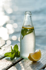 Frosted Glass Bottle with Lemon Slice and Mint Leaves on Sunny Lakeside Dock with Sparkling Water Droplets