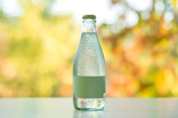 Chilled Clear Glass Bottle with Blank Label and Condensation Set Against Vibrant Yellow and Orange Blurred Outdoor Background