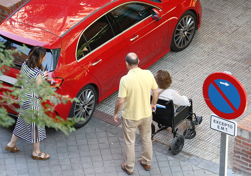 Familia con abuela en silla de ruedas y movilidad reducida con coche rojo en la calle y se&ntilde;al de tr&aacute;fico