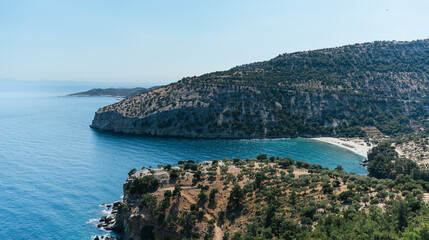 Lush vegetation enveloping steep cliffs surrounds a picturesque bay with crystal-clear turquoise water gently lapping against a secluded sandy beach in Thasos island, Greece