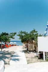 White wooden path leading to a sandy beach with sunbeds, umbrellas, straw beach huts, a Greek flag waving in the wind, and lush green trees under a clear blue sky. Thasos island. Greece.