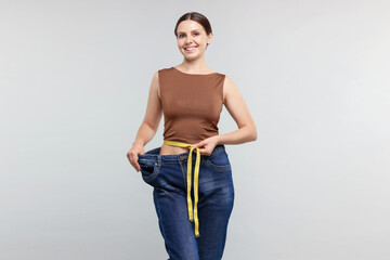 Beautiful woman with measuring tape wearing old jeans after weight loss on light grey background