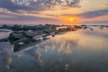 Sunset over the ocean with stones in the foreground and calm waves, creating a scenic seascape.
