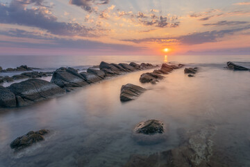 Sunset over the ocean with stones in the foreground and calm waves, creating a scenic seascape.