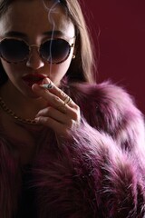 Beautiful woman in fur coat smoking cigarette on dark pink background, closeup