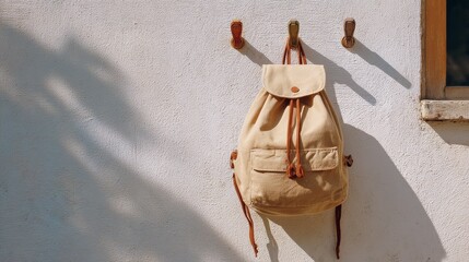 Beige canvas backpack hanging on white wall in sunlit outdoor scene