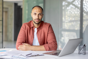 A thoughtful man sits at a desk, looking directly at the camera, with a laptop and documents nearby.